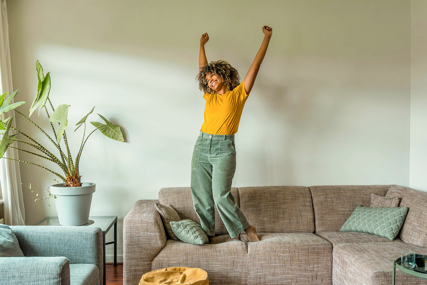 Eine junge Frau hüpft ausgelassen auf der Couch in ihrem Wohnzimmer. 