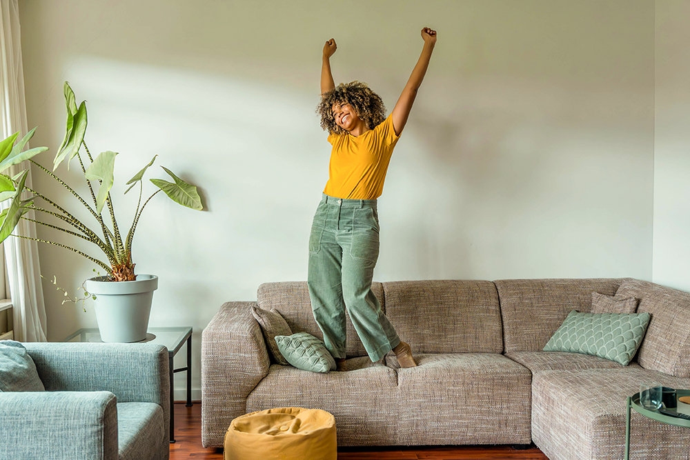 Eine junge Frau hüpft ausgelassen auf der Couch in ihrem Wohnzimmer. 