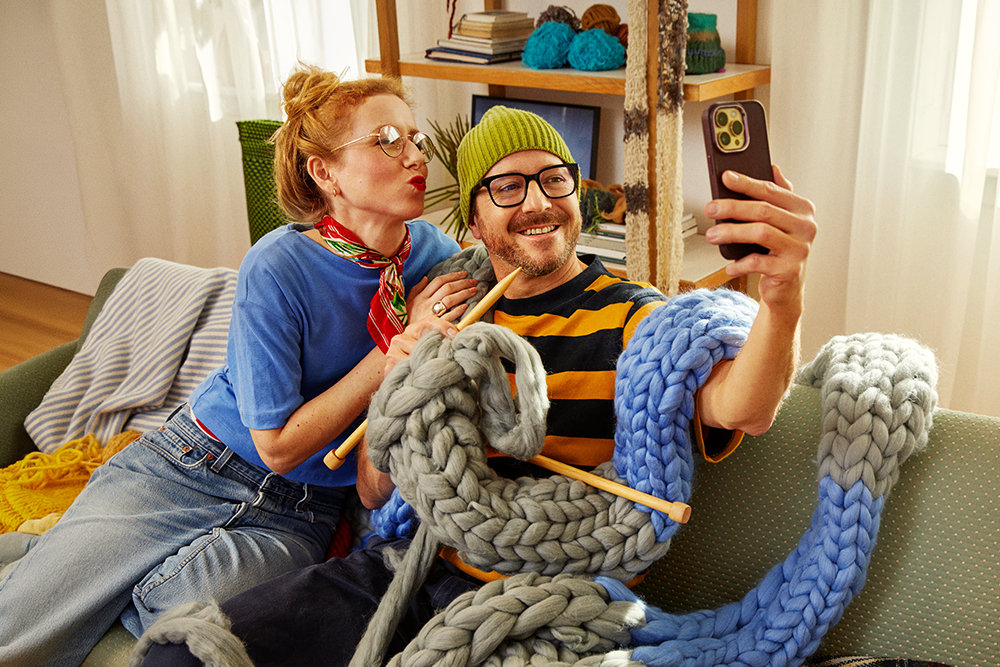 Ein Mann und eine Frau sitzen mit einem überdimensional langen, gestrickten Schaal auf einem Sofa und machen ein Selfie von sich.