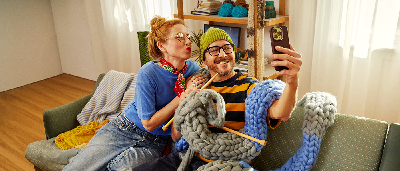 Ein Mann und eine Frau sitzen mit einem überdimensional langen, gestrickten Schaal auf einem Sofa und machen ein Selfie von sich.