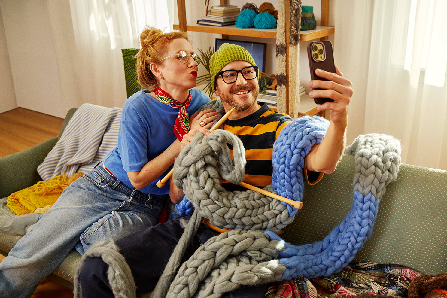 Ein Mann und eine Frau sitzen mit einem überdimensional langen, gestrickten Schaal auf einem Sofa und machen ein Selfie von sich.