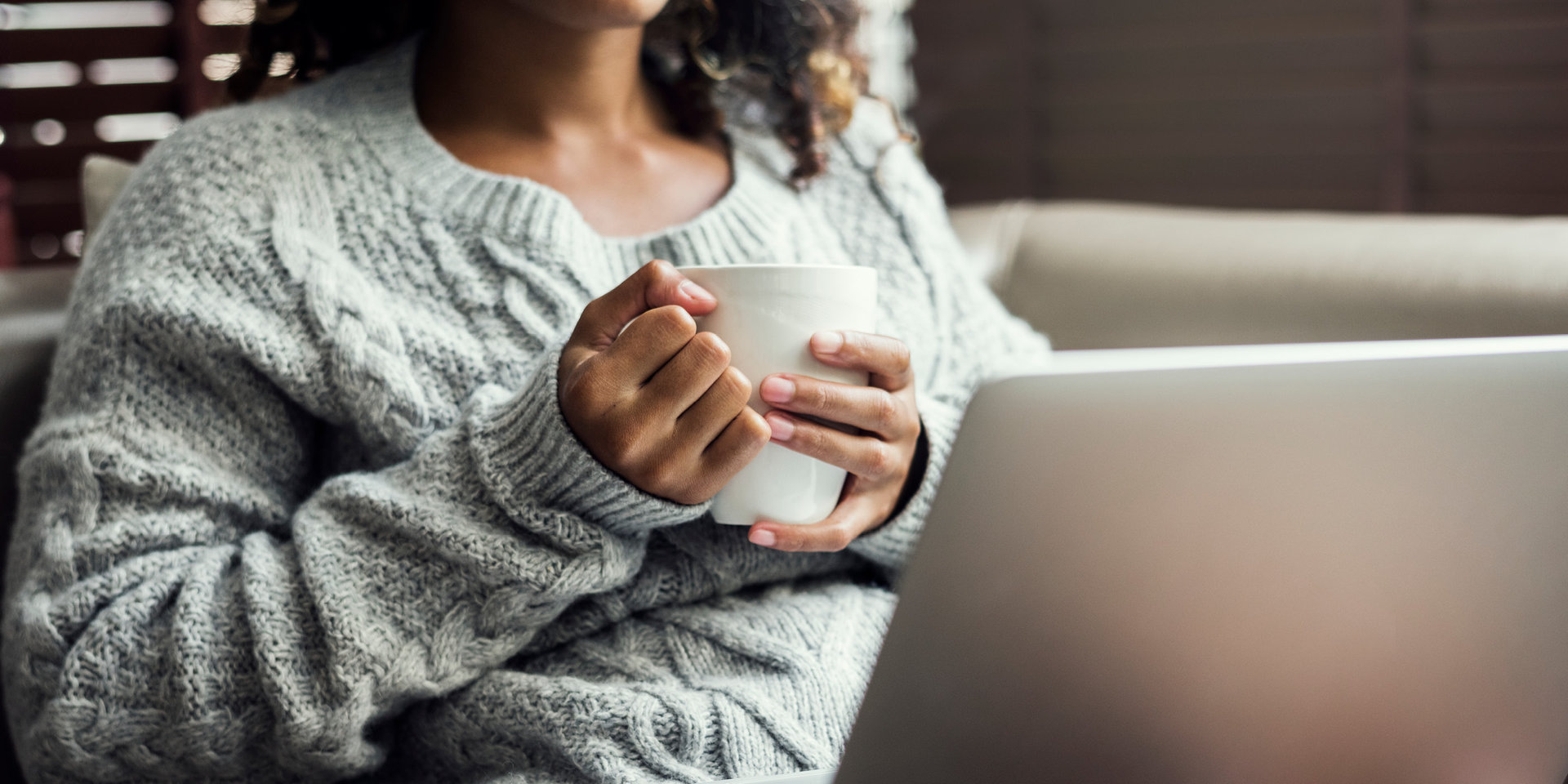 Eine Frau sitzt mit ihrem Laptop auf einem Sofa und hält eine Tasse in ihren Händen.