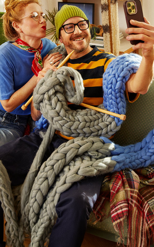 Ein Mann und eine Frau sitzen mit einem überdimensional langen, gestrickten Schaal auf einem Sofa und machen ein Selfie von sich.