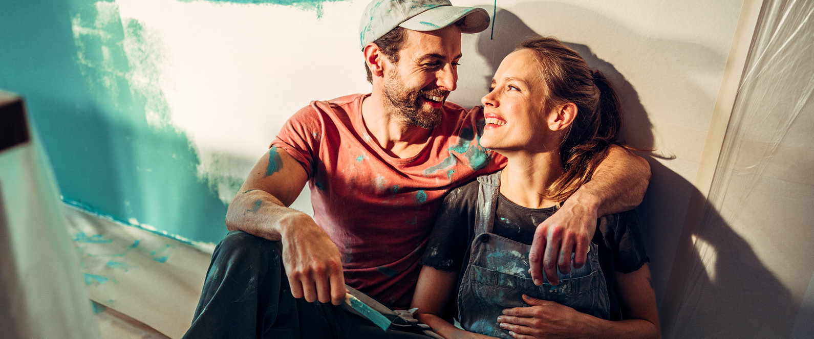 Junges Paar sitzt in Arbeitskleidung und Pinsel in der Hand vor einer frisch angestrichenen Wand und beide lächeln zufrieden.