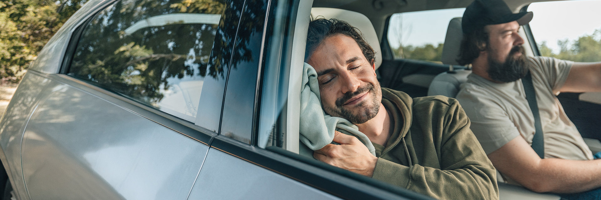 Zwei Männer in einem Auto. Der Fahrer trägt eine Kappe und ist auf den Verkehr konzentriert. Der Beifahrer schläft entspannt und lächelt dabei.