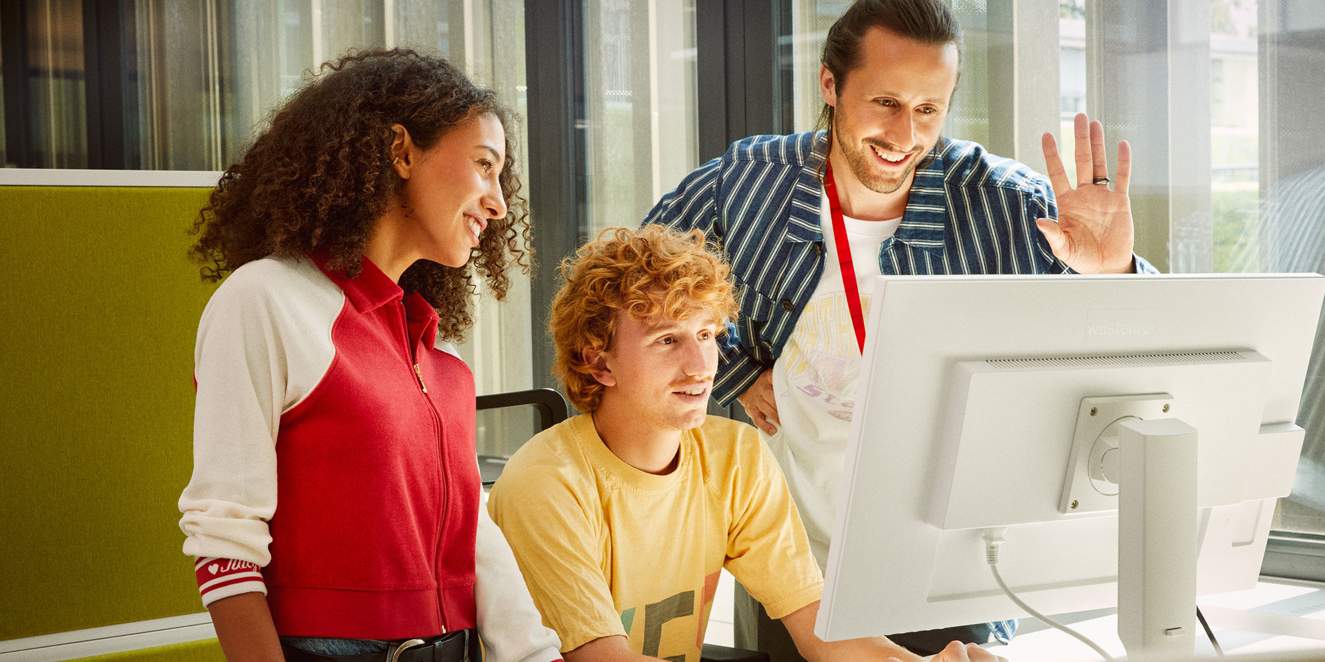 zwei Kollegen und eine Kollegin vor einem Bildschirm, während sie an einer Videokonferenz teilnehmen. Ein Kollege winkt in die Bildschirmkamera.