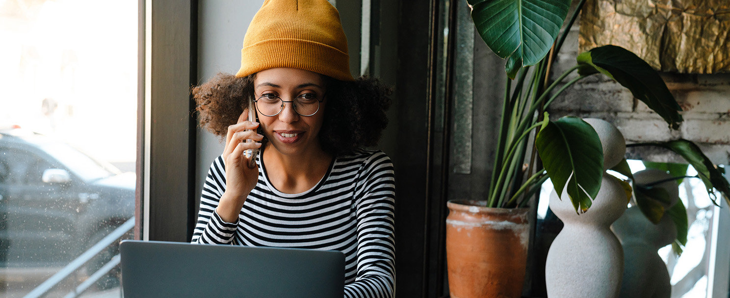 Eine Frau mit Haube und Brille sitzt vor einem Laptop und telefoniert mit dem Handy.