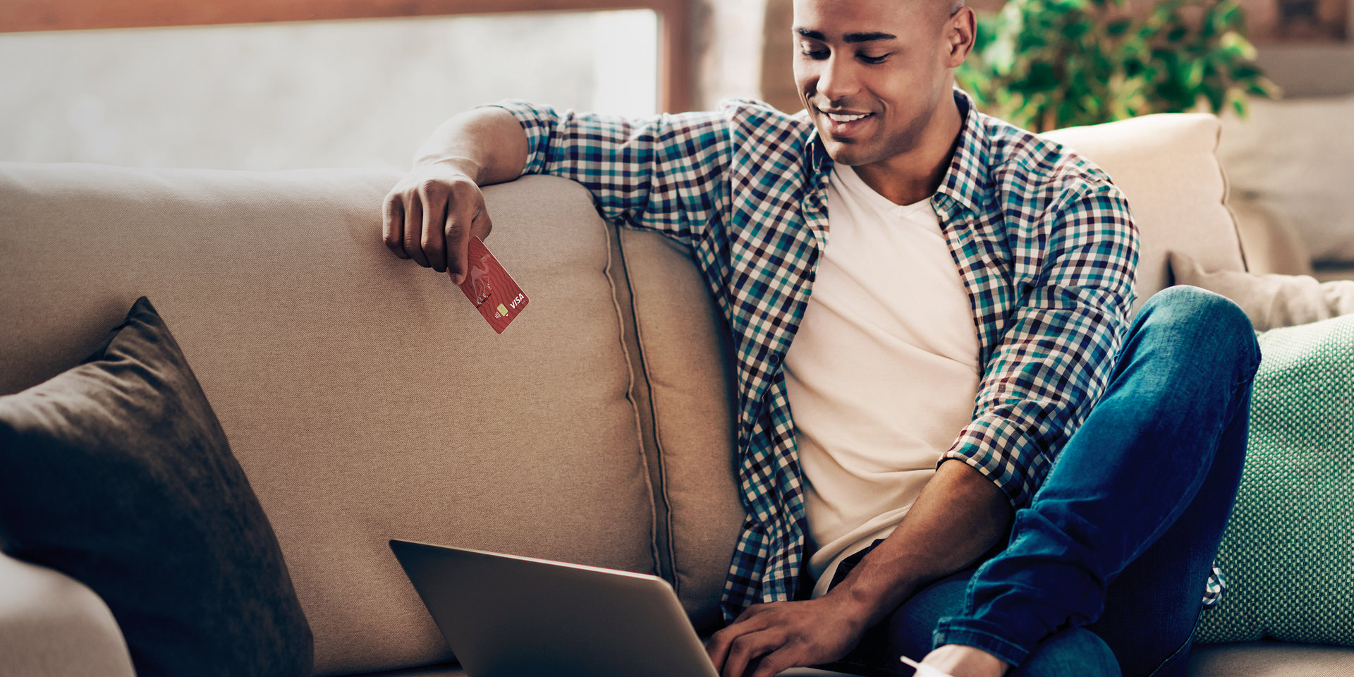 Ein Mann sitzt vor seinem Laptop auf einer Couch und hält die Debitkarte seines Wüstenrot Girokontos in der Hand.