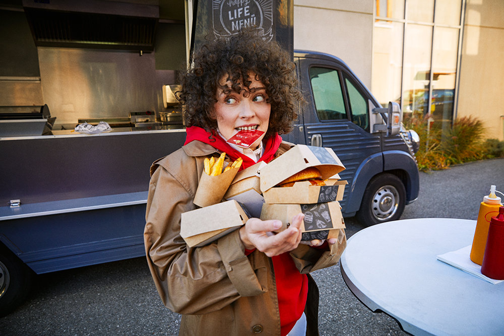 Eine junge Frau vor einem Foodtruck trägt mehrere Take-away-Boxen und hält ihre Wüstenrot Bankkarte zwischen den Zähnen. 