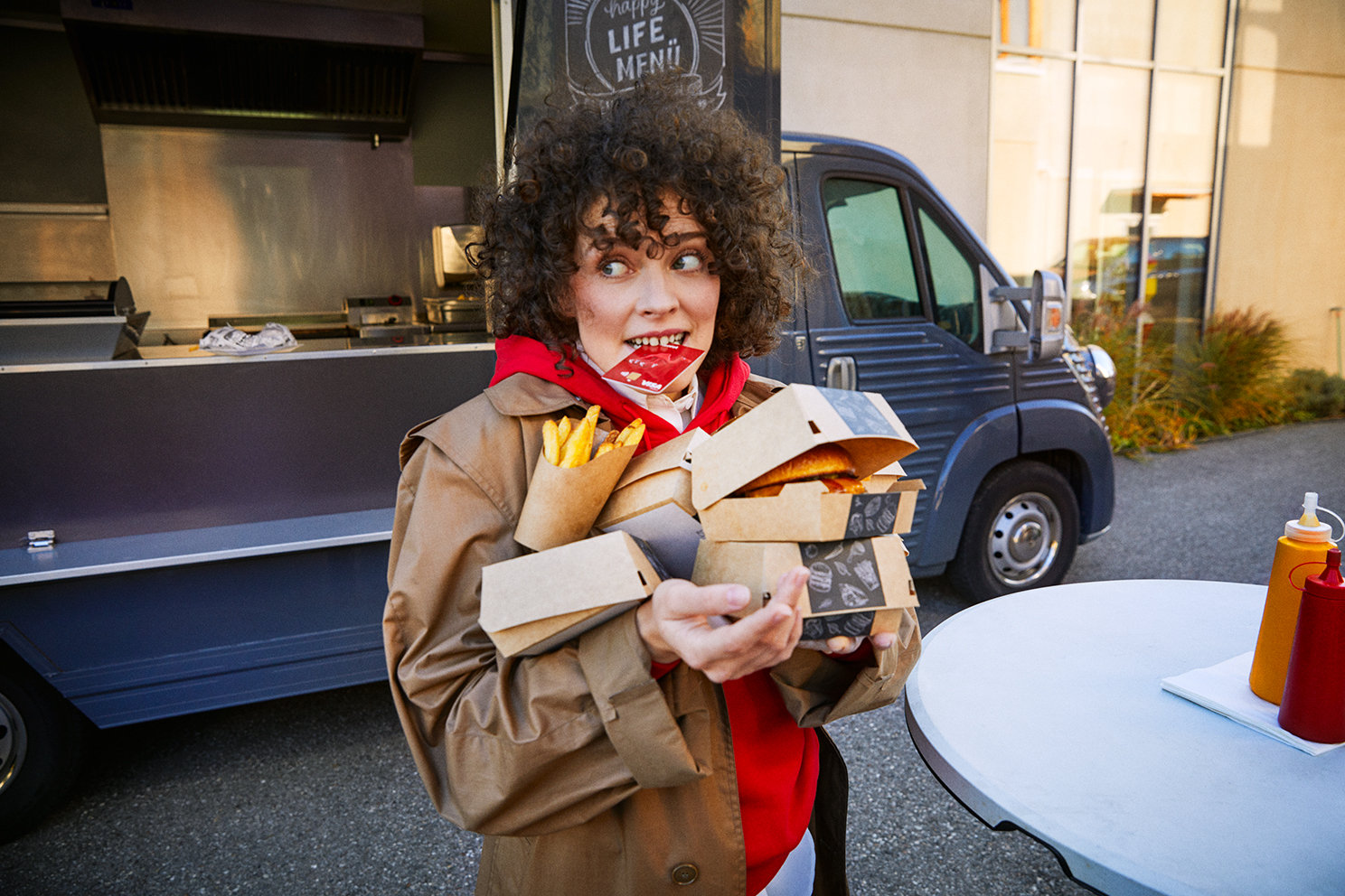Eine junge Frau vor einem Foodtruck trägt mehrere Take-away-Boxen und hält ihre Wüstenrot Bankkarte zwischen den Zähnen. 