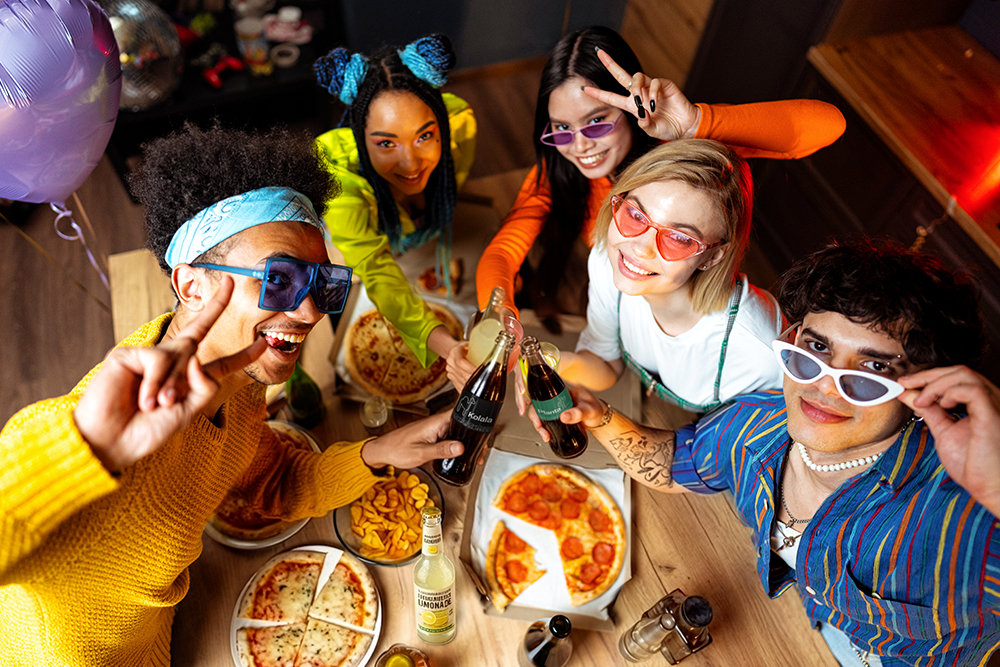 Eine Gruppe junger Leute rund um einen Tisch mit Pizza und Cola. 