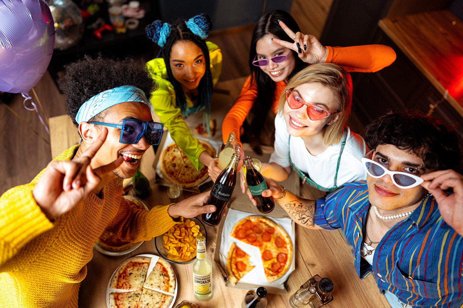 Eine Gruppe junger Leute rund um einen Tisch mit Pizza und Cola. 