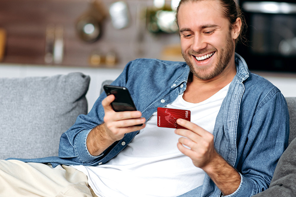Ein Mann auf einer Couch blickt lächelnd auf sein Handy und die Bankkarte zu seinem Wüstenrot Girokonto. 
