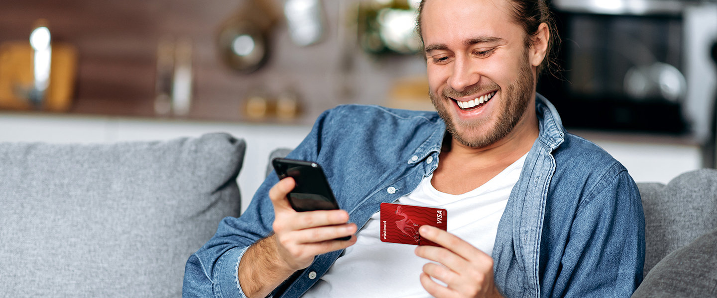 Ein Mann auf einer Couch blickt lächelnd auf sein Handy und die Bankkarte zu seinem Wüstenrot Girokonto. 
