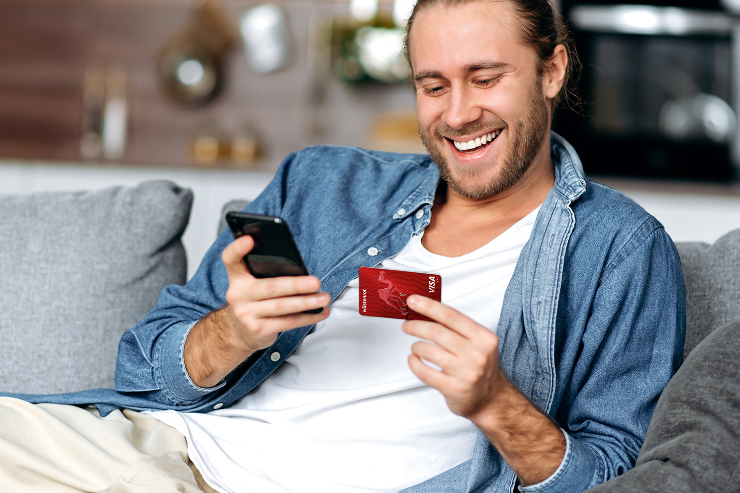 Ein Mann auf einer Couch blickt lächelnd auf sein Handy und die Bankkarte zu seinem Wüstenrot Girokonto. 