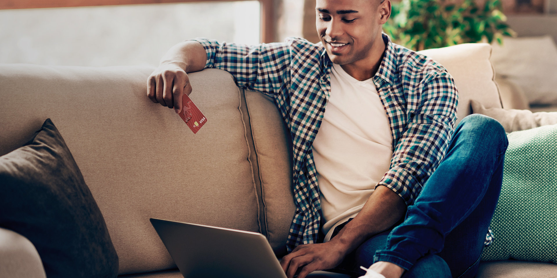 Ein Mann sitzt vor seinem Laptop auf einer Couch und hält die Debitkarte seines Wüstenrot Girokontos in der Hand. 
