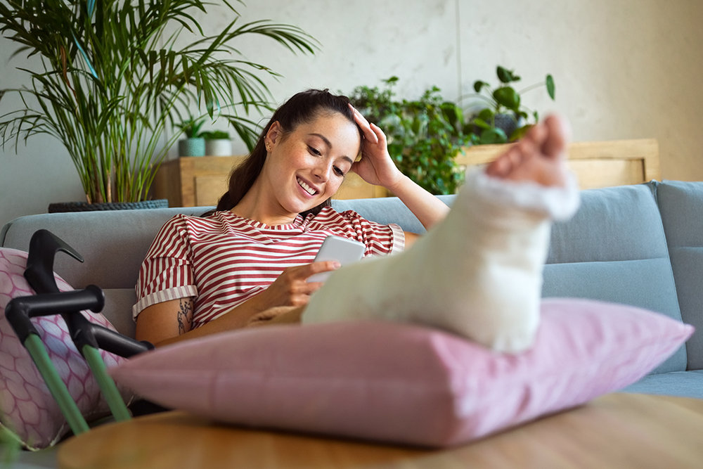 Eine lächelnde Person sitzt mit eingegipstem Bein auf einem Sofa und schaut entspannt auf ihr Smartphone. 