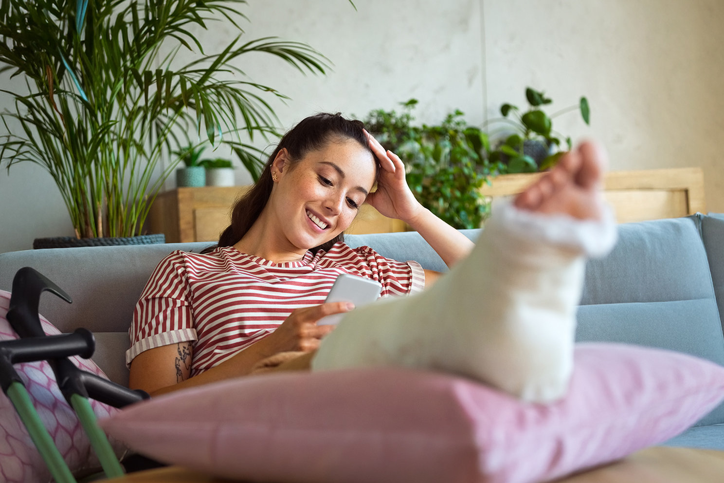Eine lächelnde Person sitzt mit eingegipstem Bein auf einem Sofa und schaut entspannt auf ihr Smartphone. 
