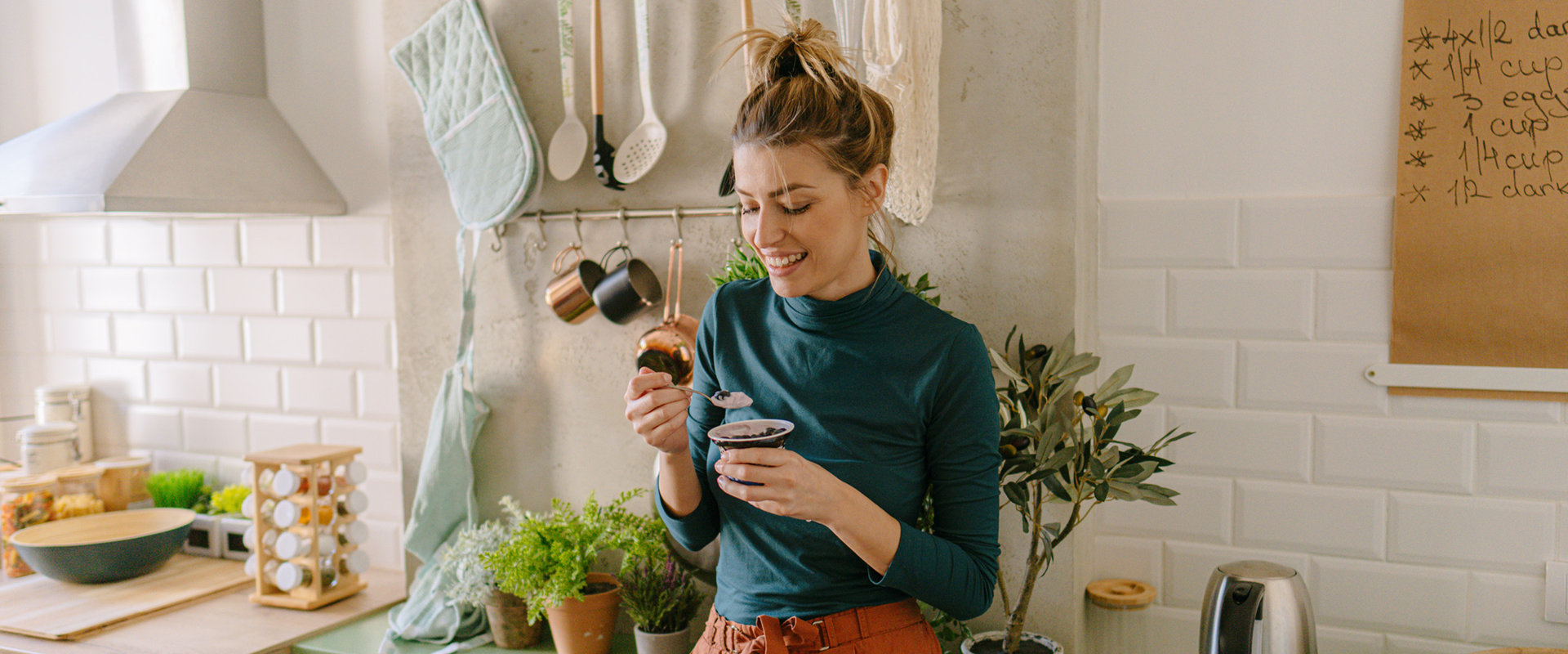 Eine Frau steht lächelnd in der Küche und isst ein Joghurt. 