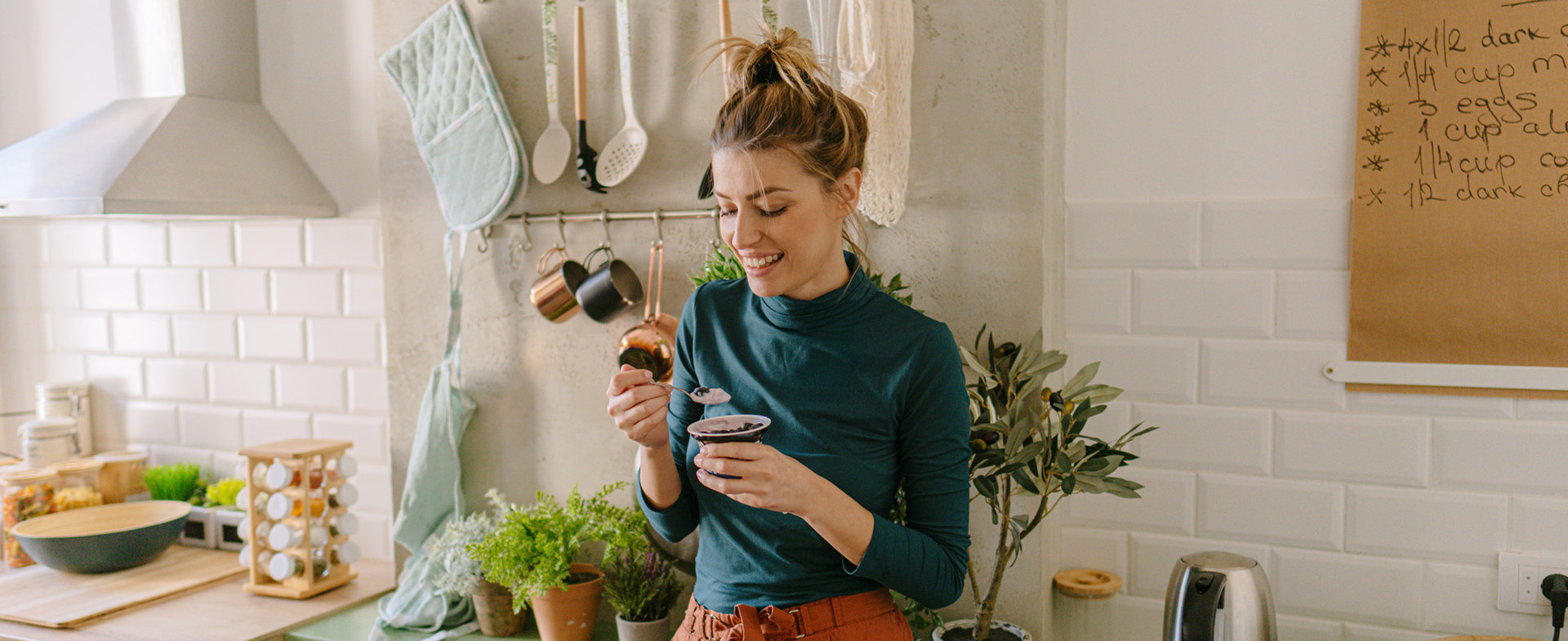 Eine Frau steht lächelnd in der Küche und isst ein Joghurt. 