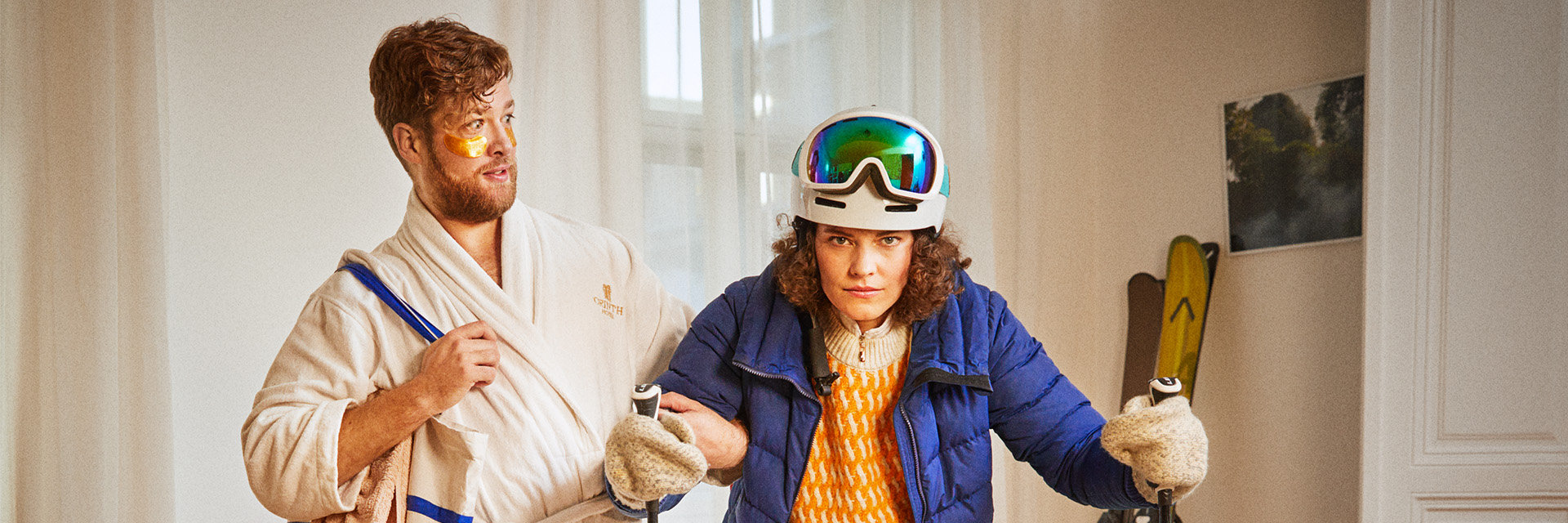 Ein Mann im Bademantel blickt verwundert zu einer Frau in Ski-Bekleidung. 