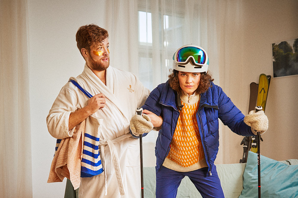 Ein Mann im Bademantel blickt verwundert zu einer Frau mit Ski-Bekleidung. 