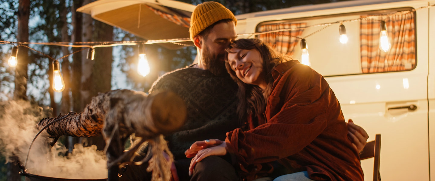 Zwei Personen sitzen vor einem Wohnwagen an einer Feuerstelle.