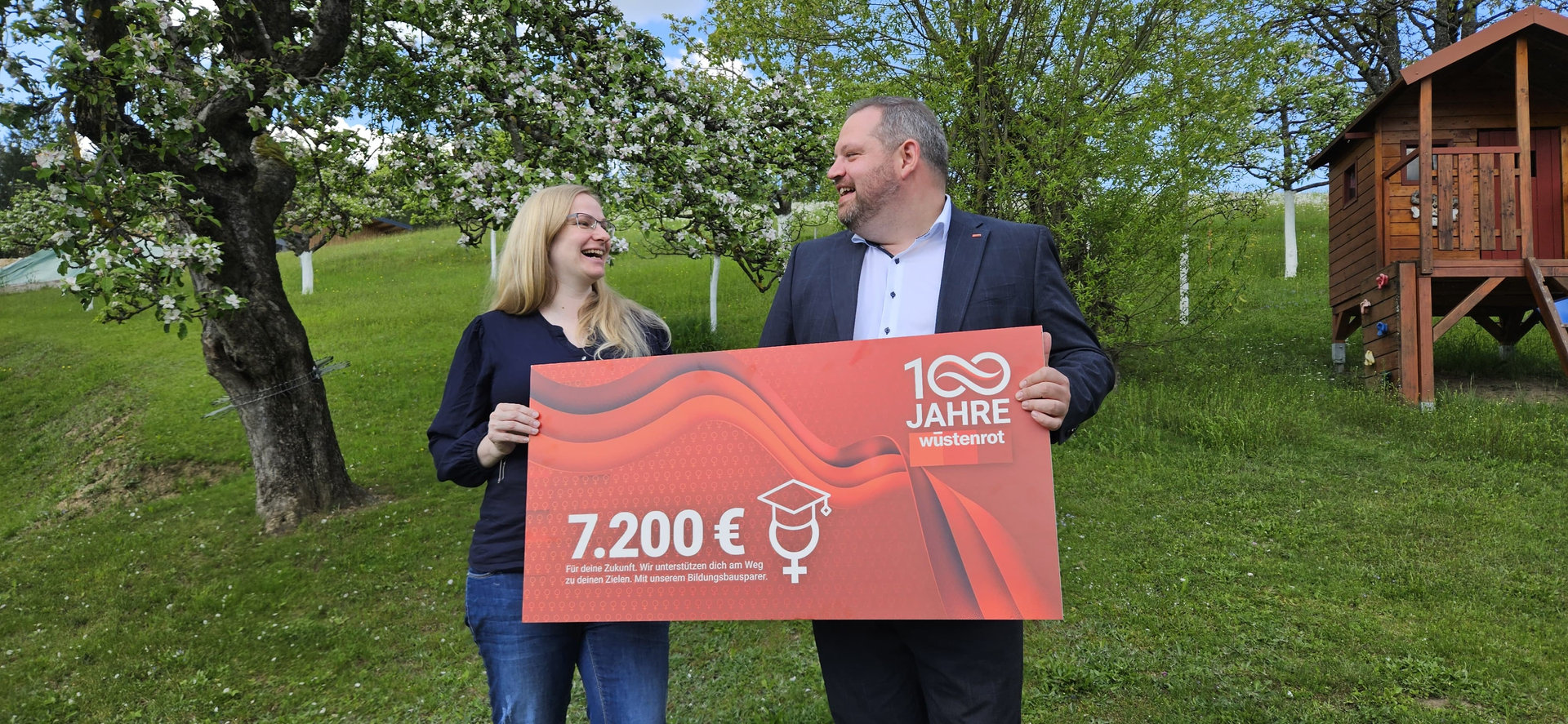 Blonde Frau und ein Mann halten einen Wüstenrot Scheck über 7.200 Euro in der Hand und lachen sich an