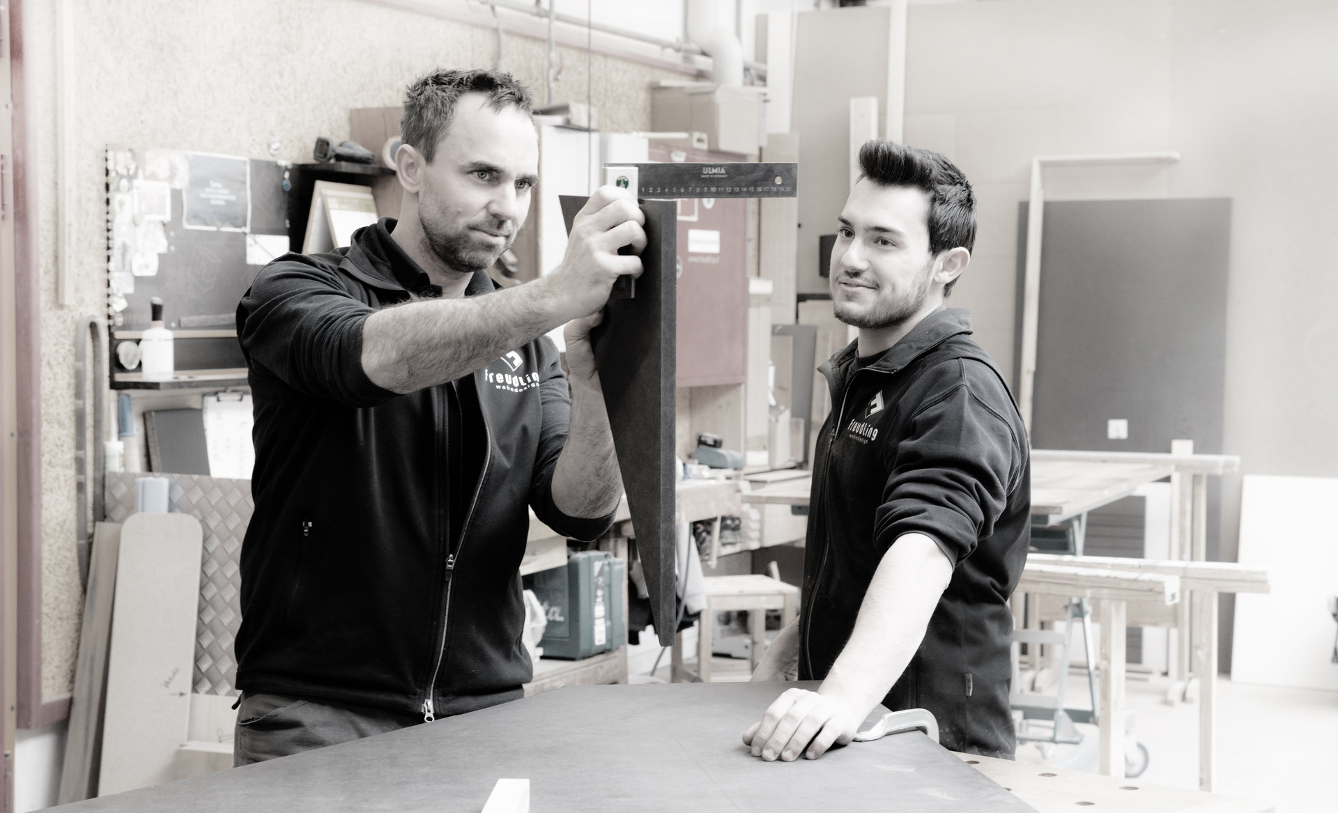 Ein Ausbildner zeigt einem Lehrling in der Werkstatt den Umgang mit einem Holzwerkzeug – Szene aus der handwerklichen Ausbildung bei Freudling Wohndesign