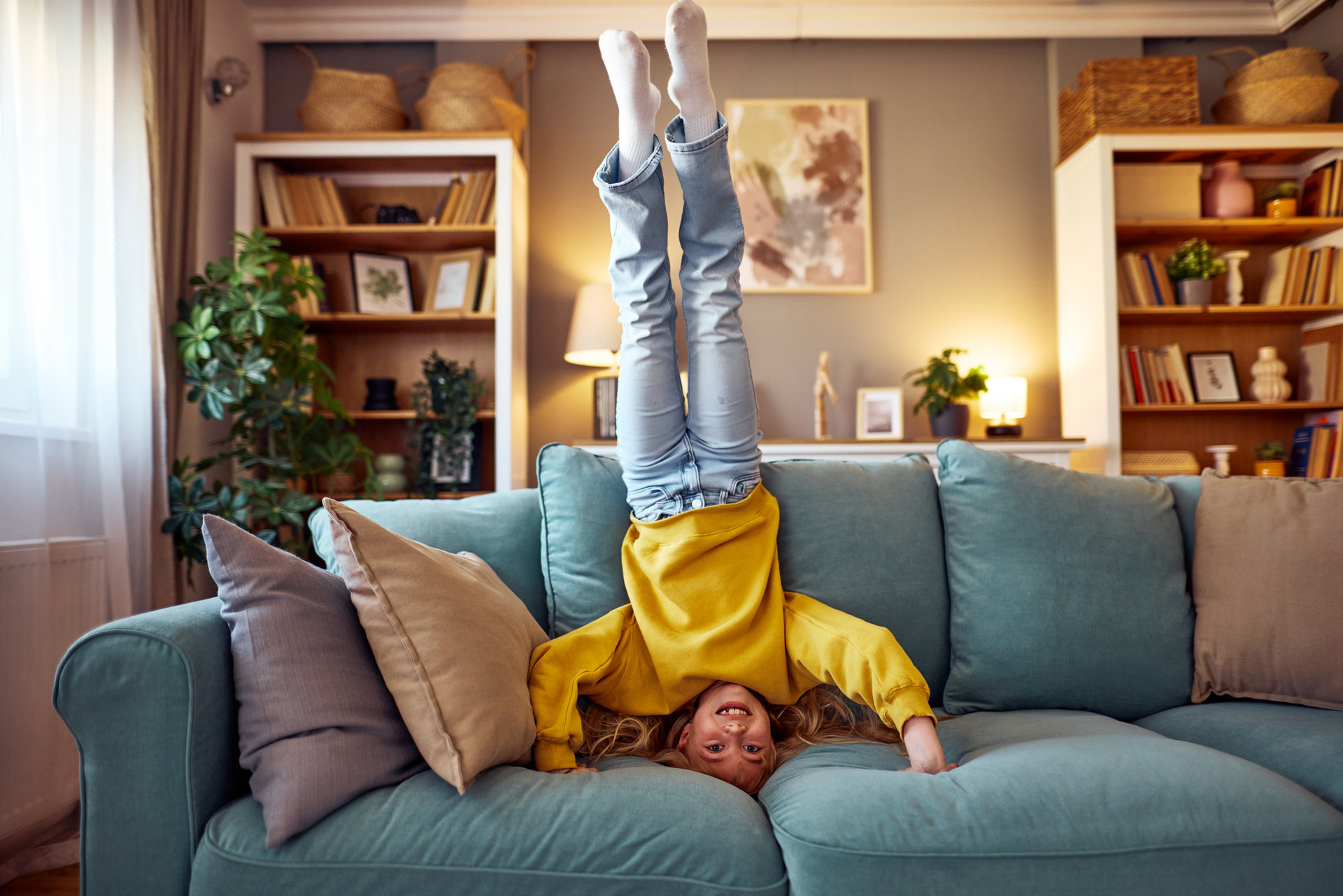 Smiling girl enjoying her leisure time, playfully doing a handstand upside down on the sofa in a cozy modern living room, radiating happiness and carefree energy at home