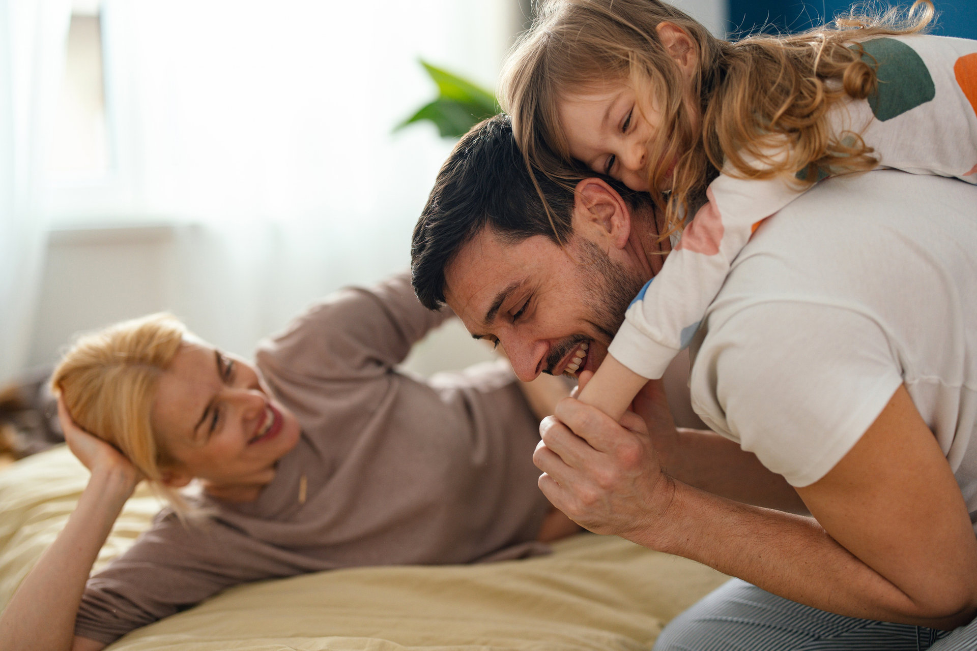 Smiling father and mother laughing and playing with their daughter while lying on a bed.