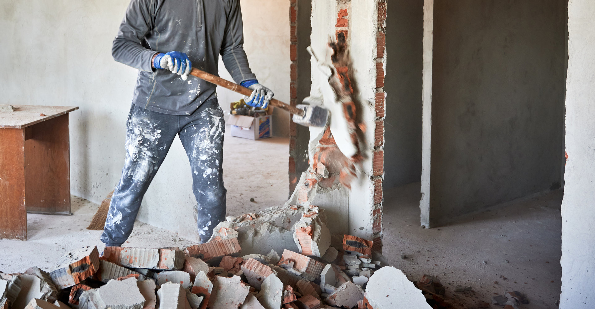 1390791404 Close up of sledgehammer blow on brick, plaster. Workman striking devastating blow at remnants of interoom wall against backdrop of table and other plastered walls.