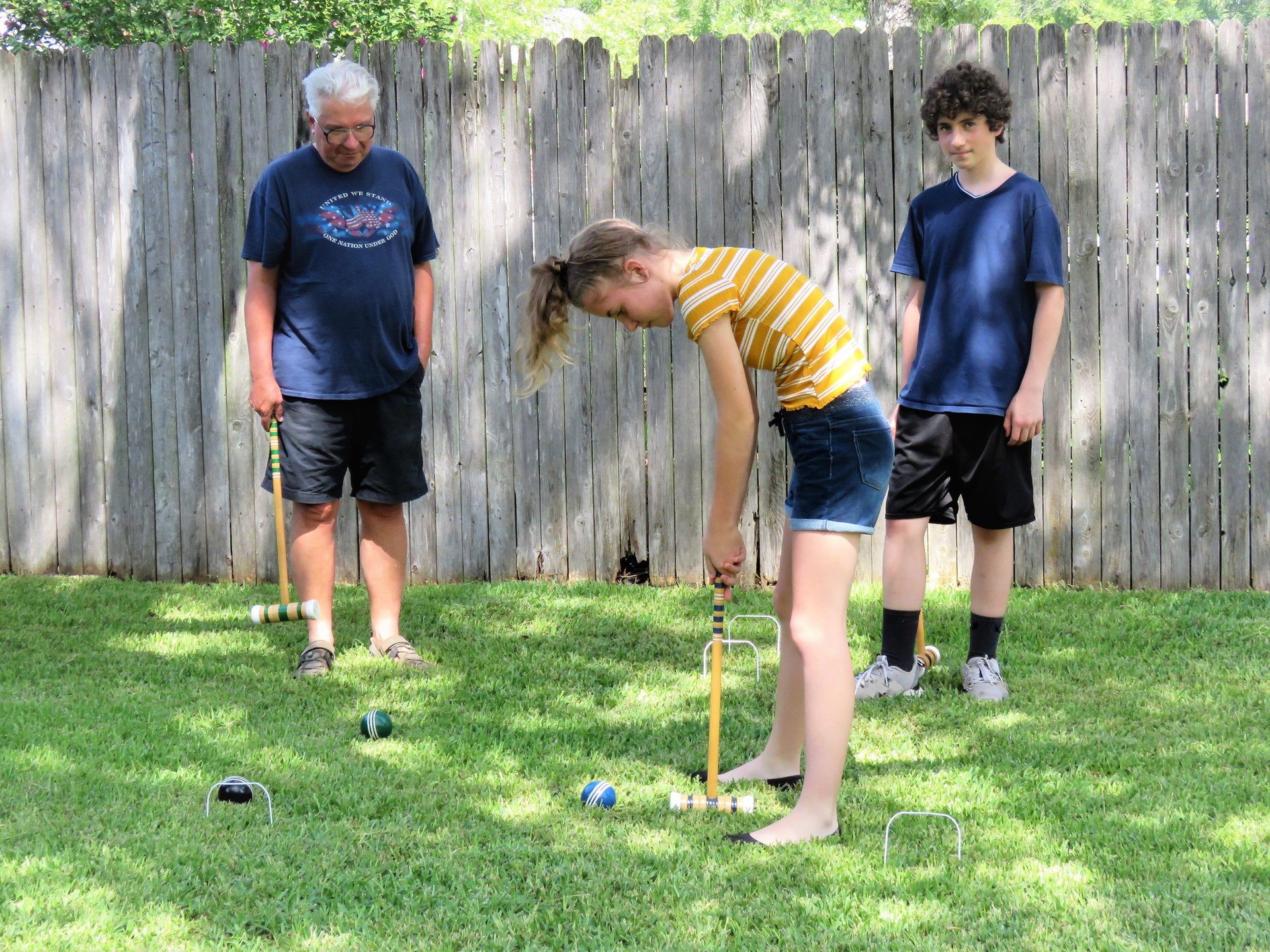 Croquet game--balls, mallot, wickets, stake--played by a grandfather and his teen grandchildren. Intergenerational. Family.