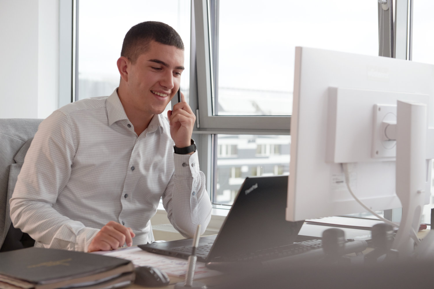 Mann im Hemd hält ein Handy an sein Ohr und sitzt vor einem Bildschirm