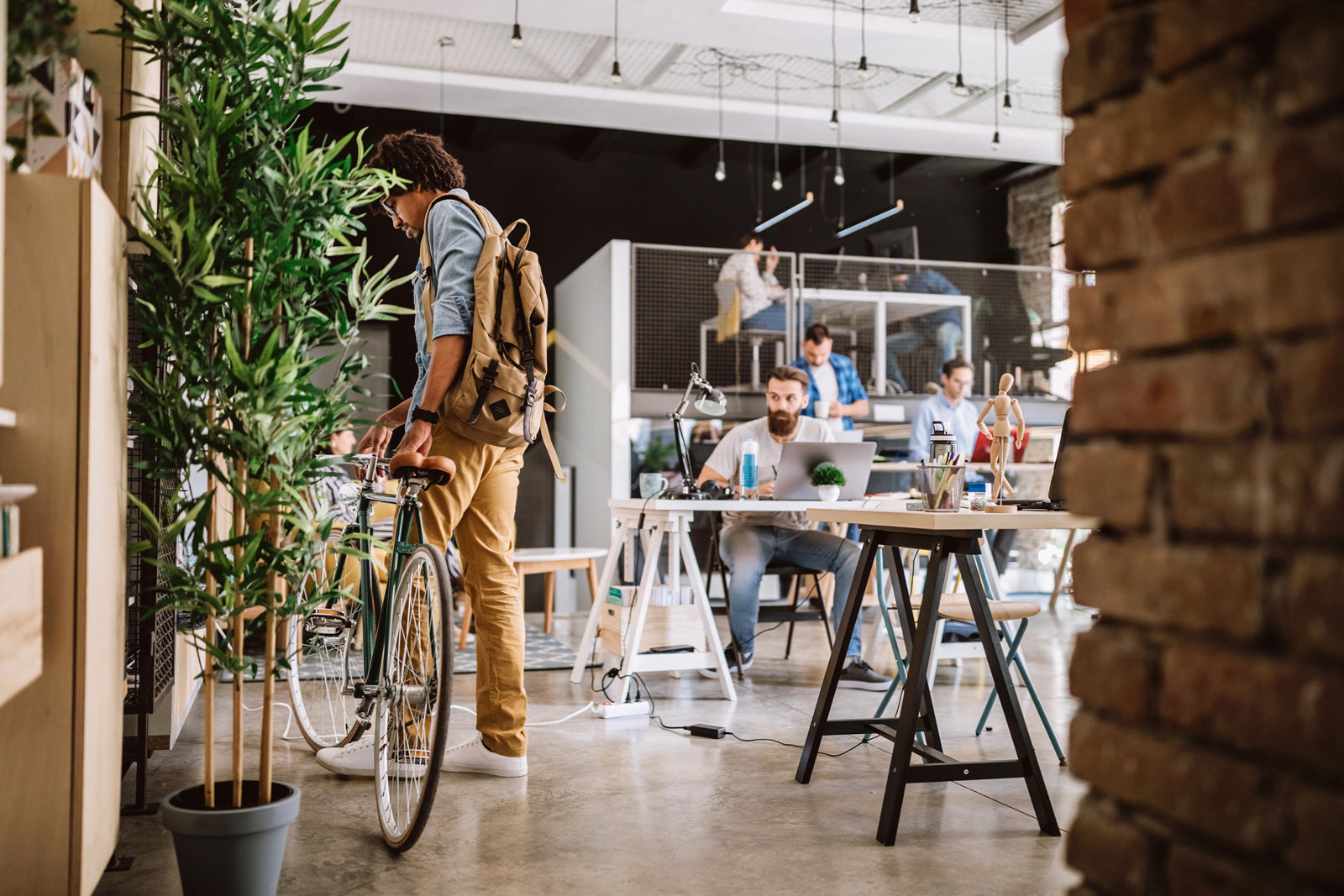 Mann mit Rucksack, der gerade sein Fahrrad im Office schiebt. Neben ihm die Büro-Kolleg:innen sitzend vor ihren Laptops.