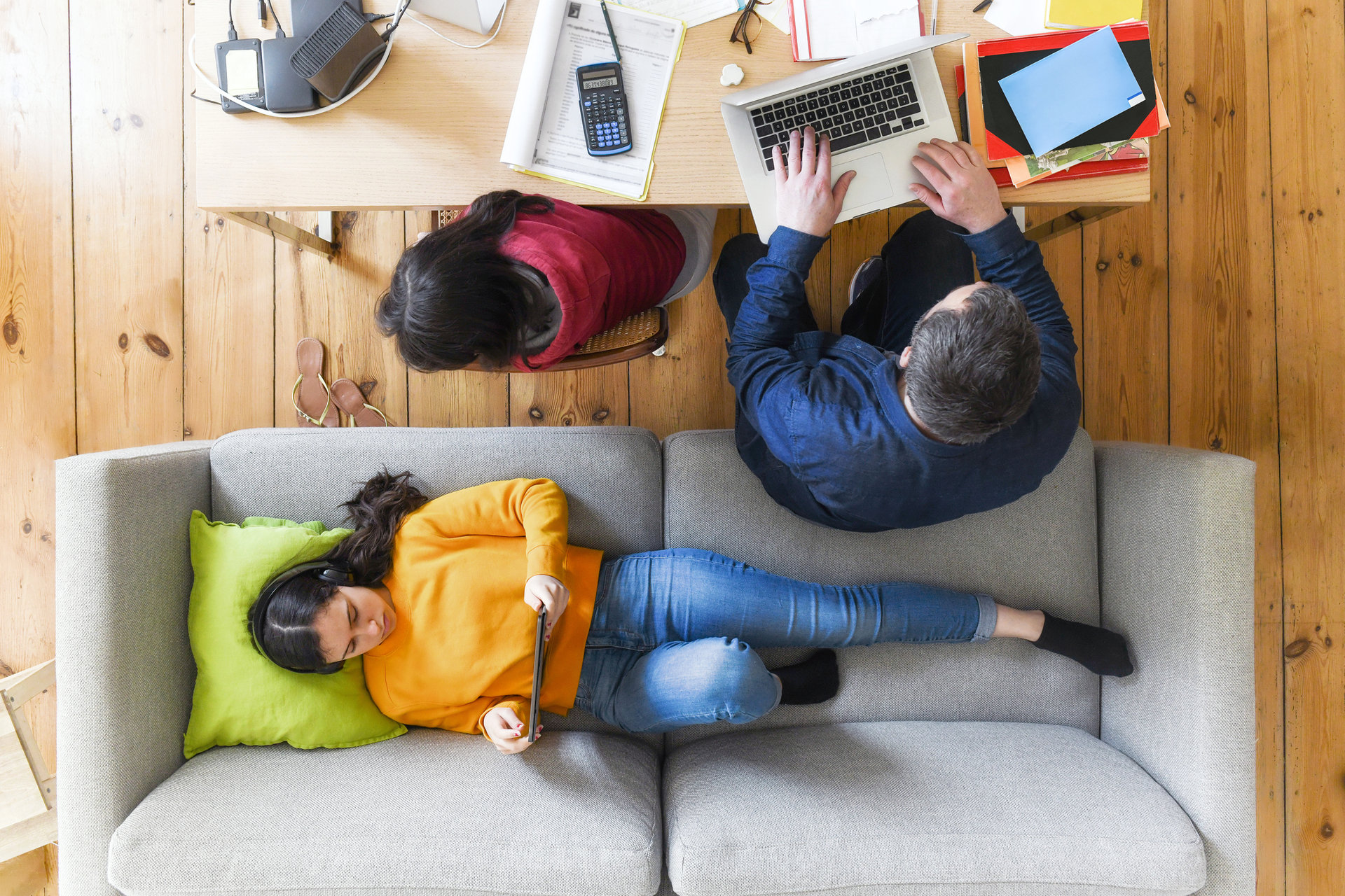 overhead view on man working at table in living room beside daughter doing home work, second daughter looking at tablet on sofa
