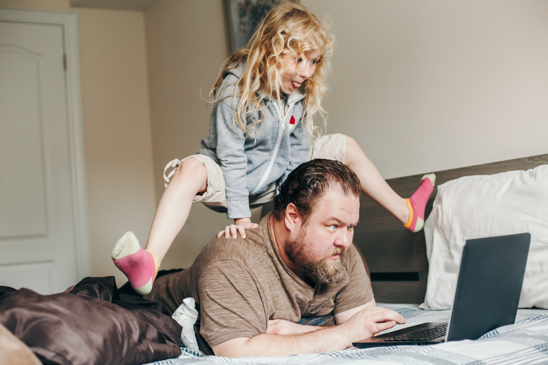 Work from home with kids children. Father working on laptop in bedroom with child daughter on his back. Funny candid family moment. New normal during coronavirus quarantine lockdown.