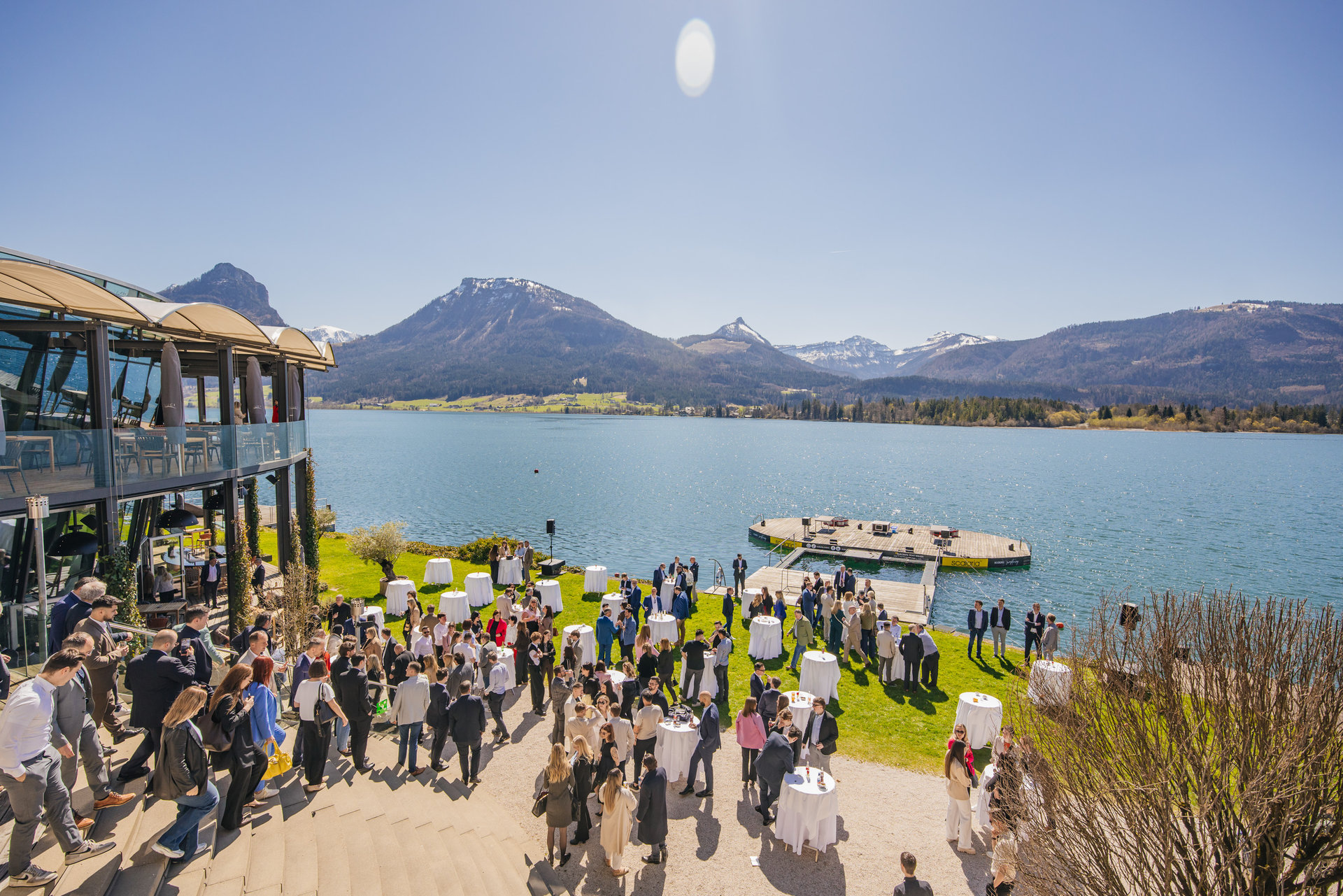 Große Personengruppe bei einer Outdoor-Veranstaltung auf einer Terrasse am See, mit Stehtischen auf einer grünen Rasenfläche, einem Steg am Wasser und einer Berglandschaft im Hintergrund bei sonnigem Wetter.
