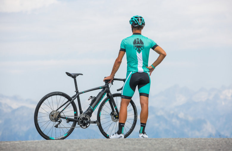 Person in türkisem Radleranzug neben schwarzem Fahrrad blickt auf die Berge