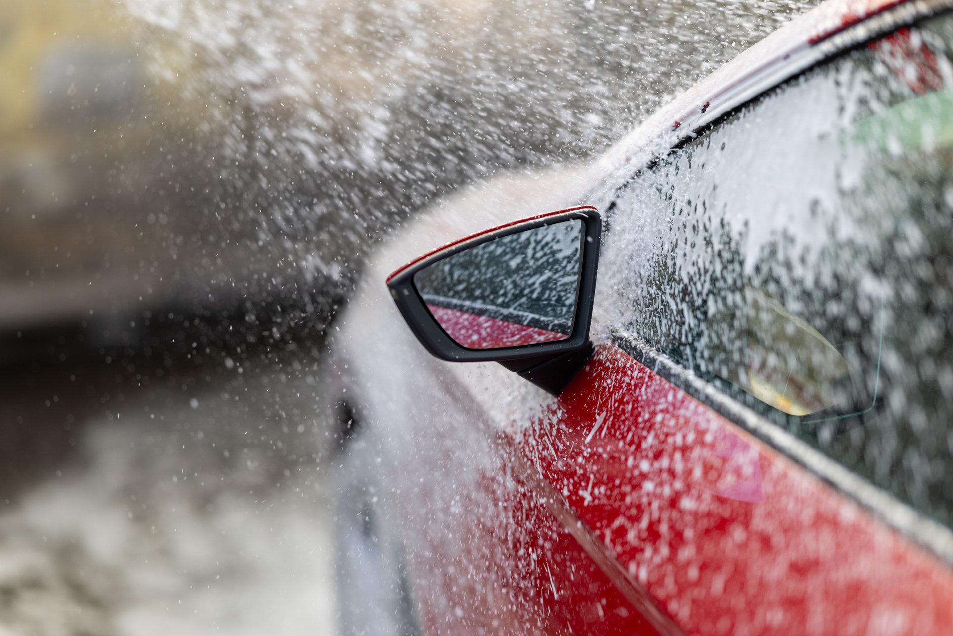A sleek red car being cleaned during an automated car wash. Water and soap create dynamic splashes over the vehicle, emphasizing cleanliness and maintenance.