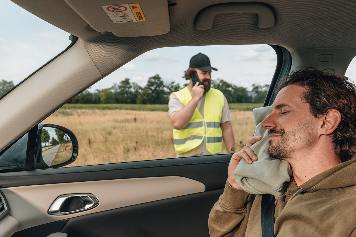 Person entspannt im Fahrzeug, während eine weitere Person außerhalb des Fahrzeugs die Pannenhilfe organisiert. 