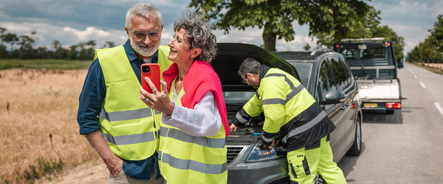 Zwei Personen stehen unbesorgt neben einem liegengebliebenen Auto am Straßenrand, während ein Pannenhelfer das Fahrzeug überprüft. 