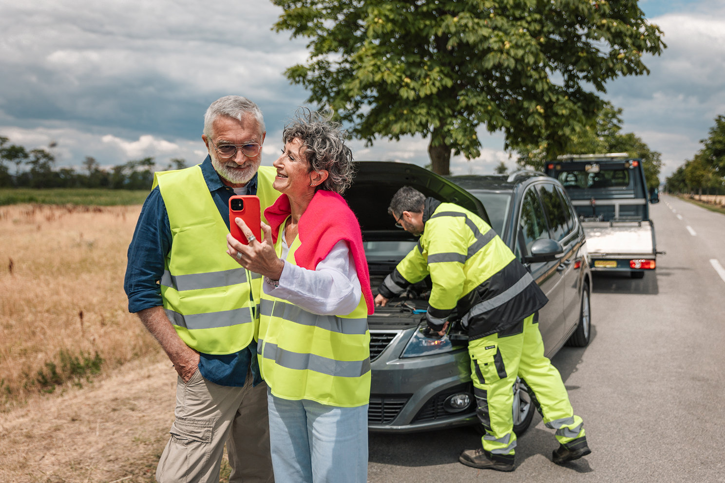 Zwei Personen stehen unbesorgt neben einem liegengebliebenen Auto am Straßenrand, während ein Pannenhelfer das Fahrzeug überprüft. 