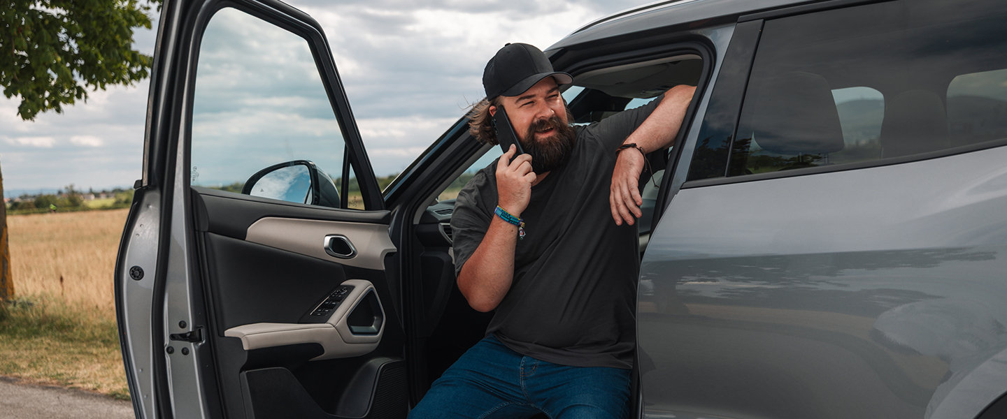 Mann mit Bart und Kappe sitzt nach einer Panne in seinem Auto bei geöffneter Tür und kontaktiert mit seinem Handy die Notfall-Hilfe.