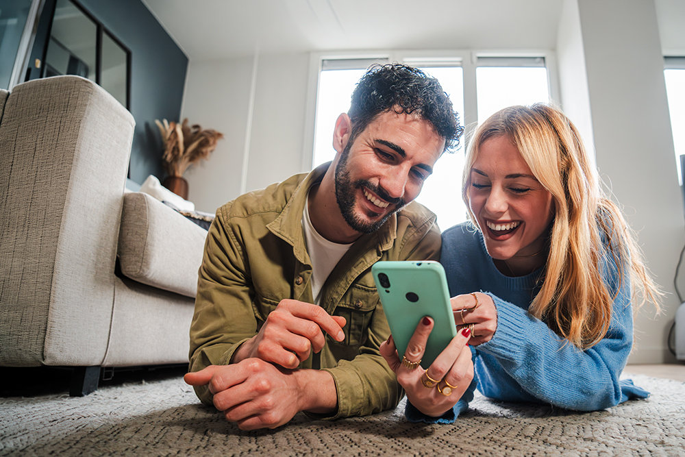 Zwei Personen liegen auf dem Boden und betrachten lächelnd gemeinsam ein Smartphone.