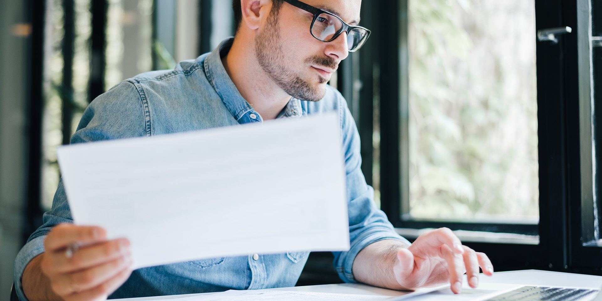 Ein Mann arbeitet an seinem Computer und hält dabei in einer Hand einen Ausdruck.