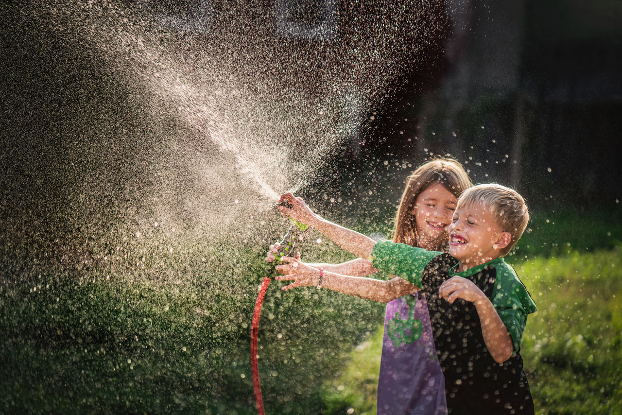 Happy little boy and girl playing with garden hose in the backyard.